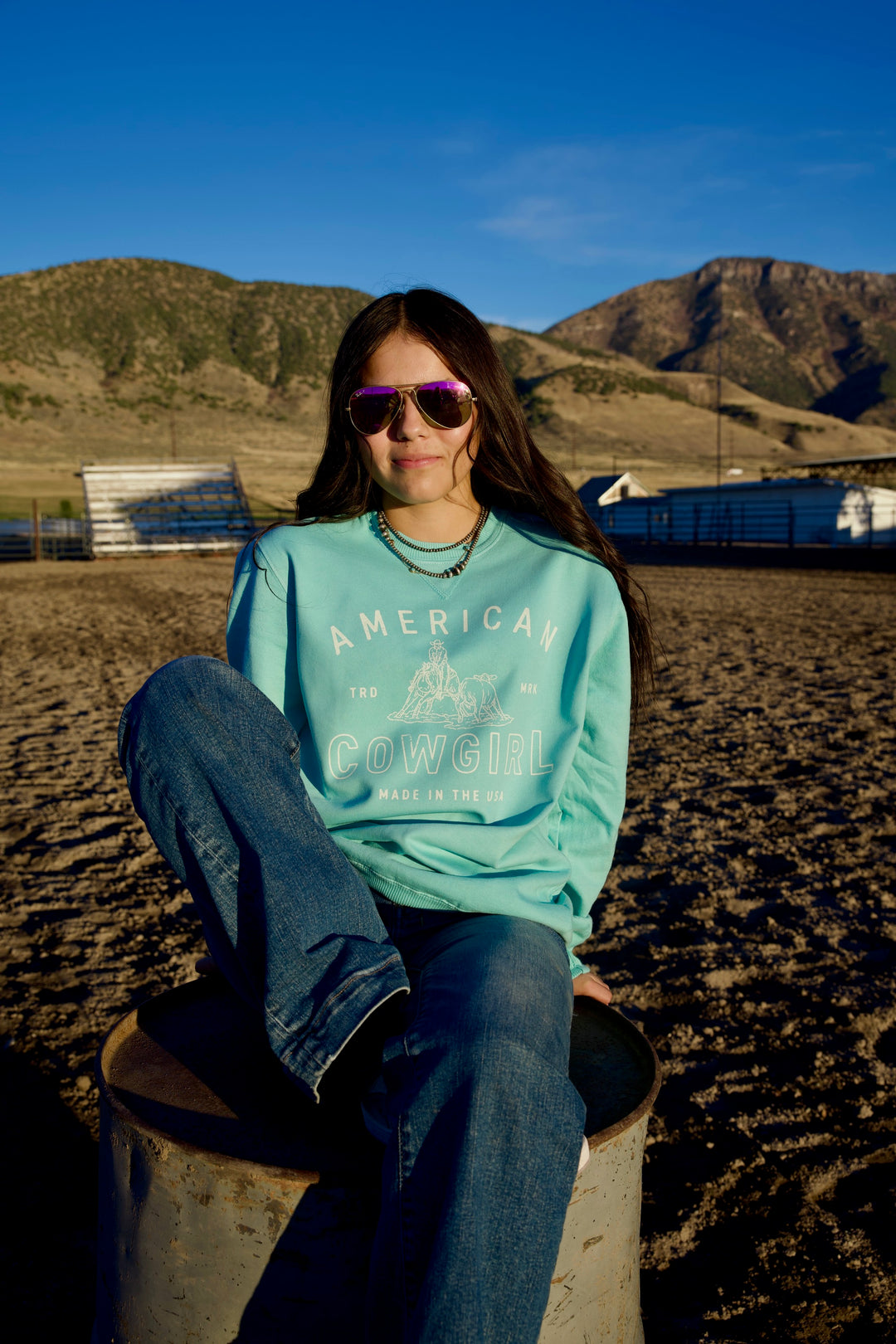 A person wearing sunglasses and a light blue crewneck/sweatshirt with the text 'AMERICAN COWGIRL' printed on the front, sitting on a metal object with a rural landscape in the background.