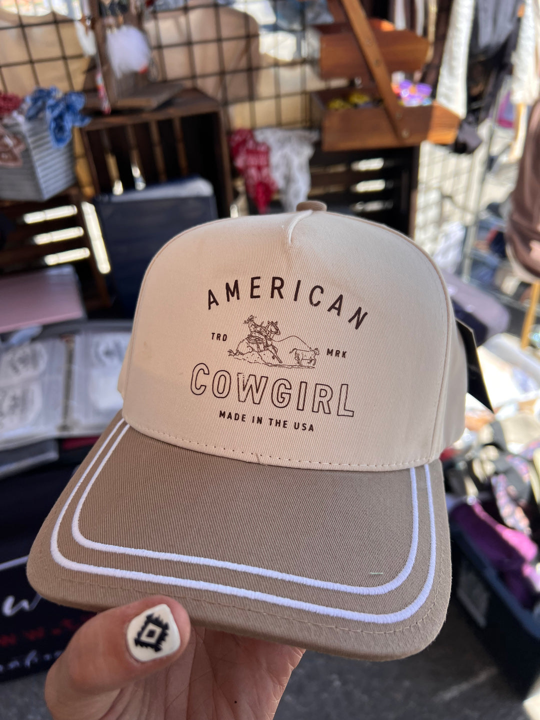 Beige cap with 'American Cowgirl' branding held by a hand in a store setting.