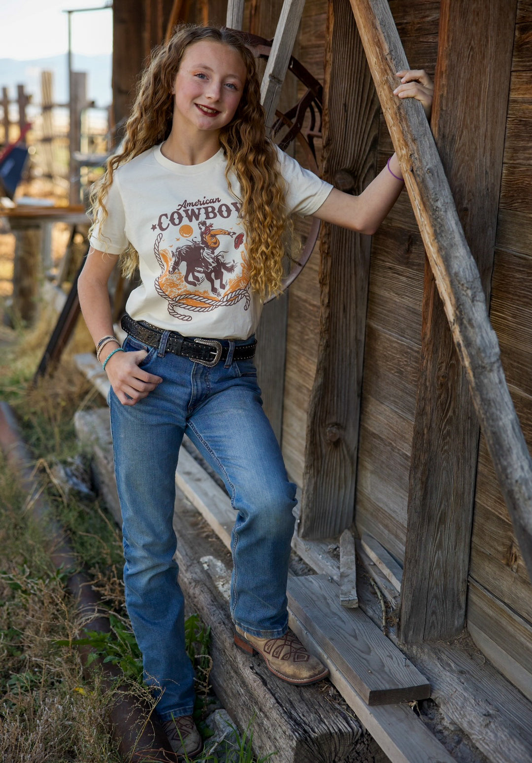 A person wearing a 'American Cowboy' graphic tee with a design of a cowboy riding a horse, paired with blue jeans and brown boots, standing near a rustic wooden structure.
