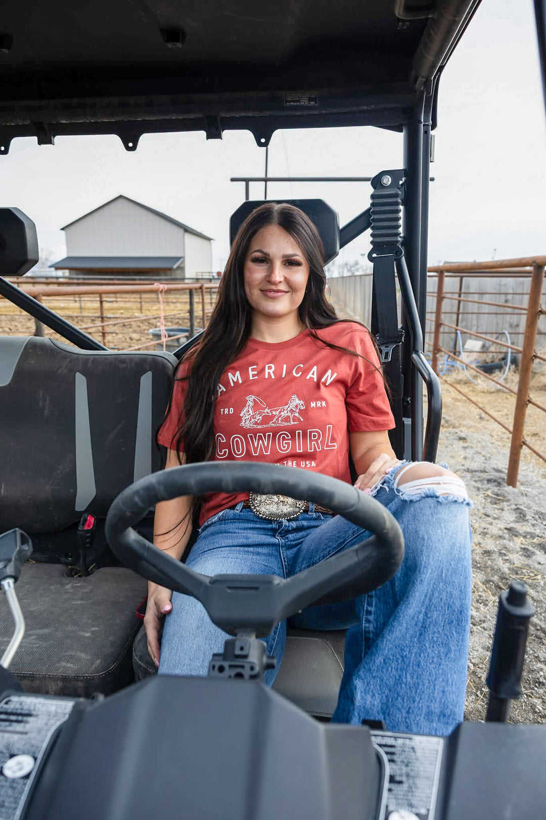 A woman sitting in a vehicle wearing a red graphic tee with the text 'American Cowgirl' and 'Made in the USA' on it, along with an image of two people team roping.