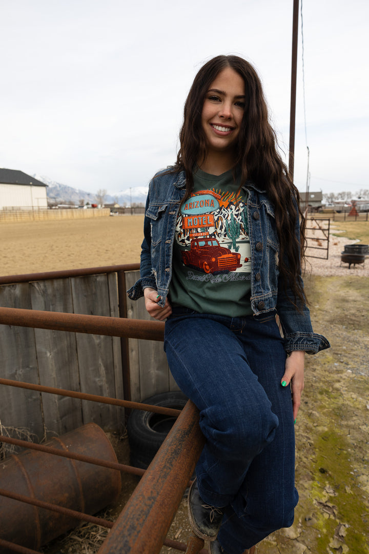 A person wearing a graphic tee with a vintage pickup truck and cacti design, sitting on a metal gate with a dessert landscape in the background.