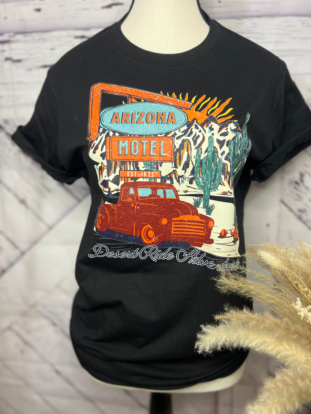 A person wearing a graphic tee with a vintage pickup truck and cacti design, sitting on a metal gate with a dessert landscape in the background.