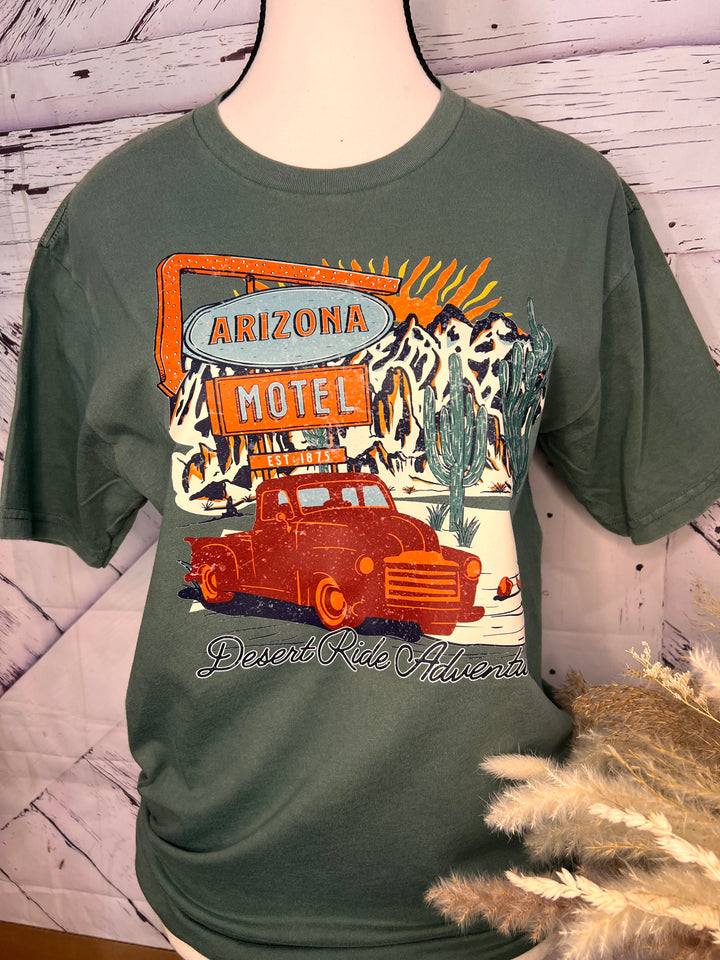 A person wearing a graphic tee with a vintage pickup truck and cacti design, sitting on a metal gate with a dessert landscape in the background.