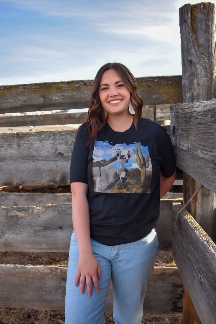 A person standing by a fence, wearing a black t-shirt with a graphic design of a donkey in the foreground.