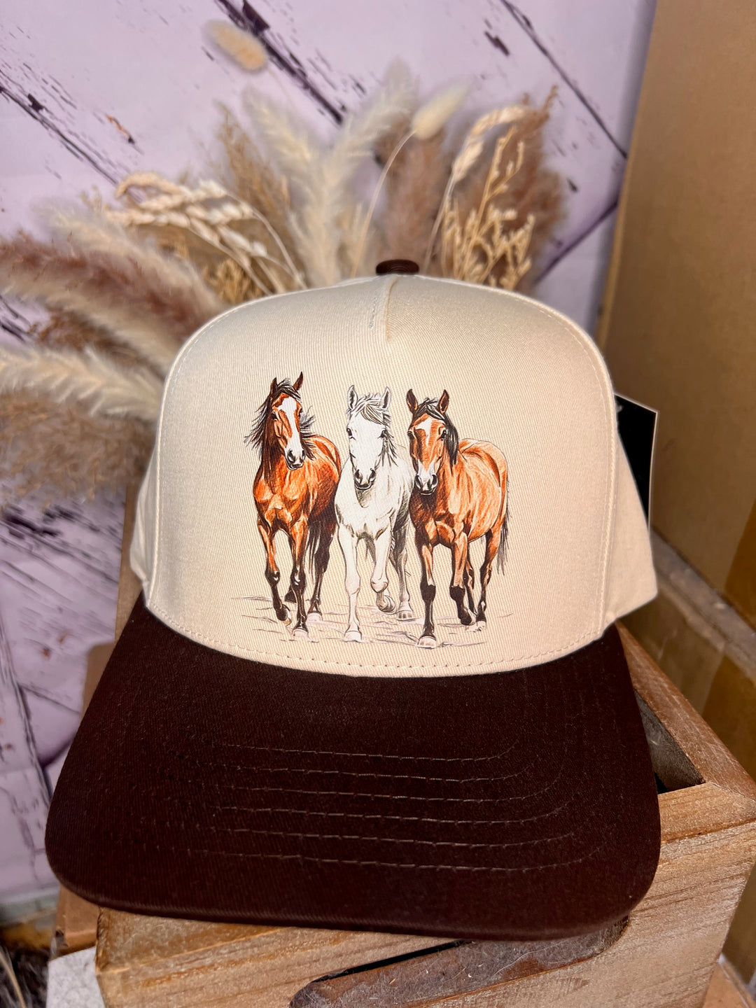 A beige and brown ballcap with a graphic of three running horses on the front and a brown bill.