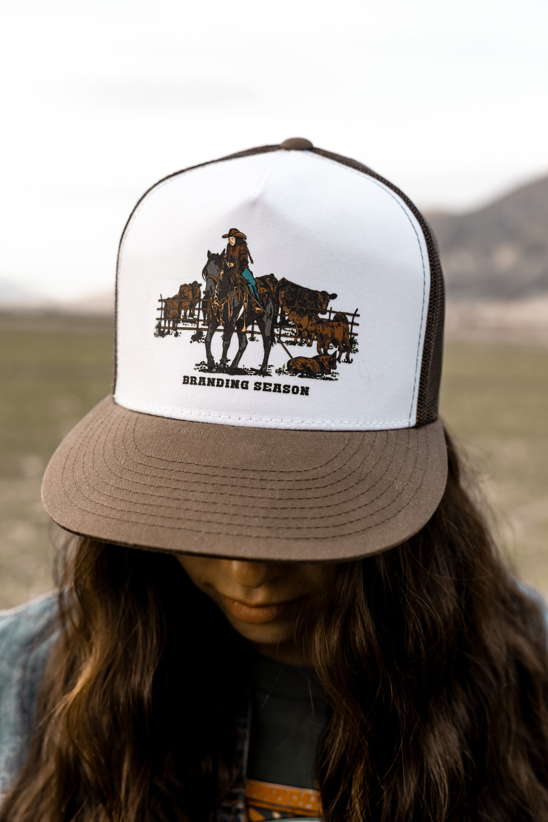 A ballcap with a graphic design of a cowgirl on a horse and text 'Branding Season'. The cap has a brown flat brim and white front with the design, while the rest is solid white.