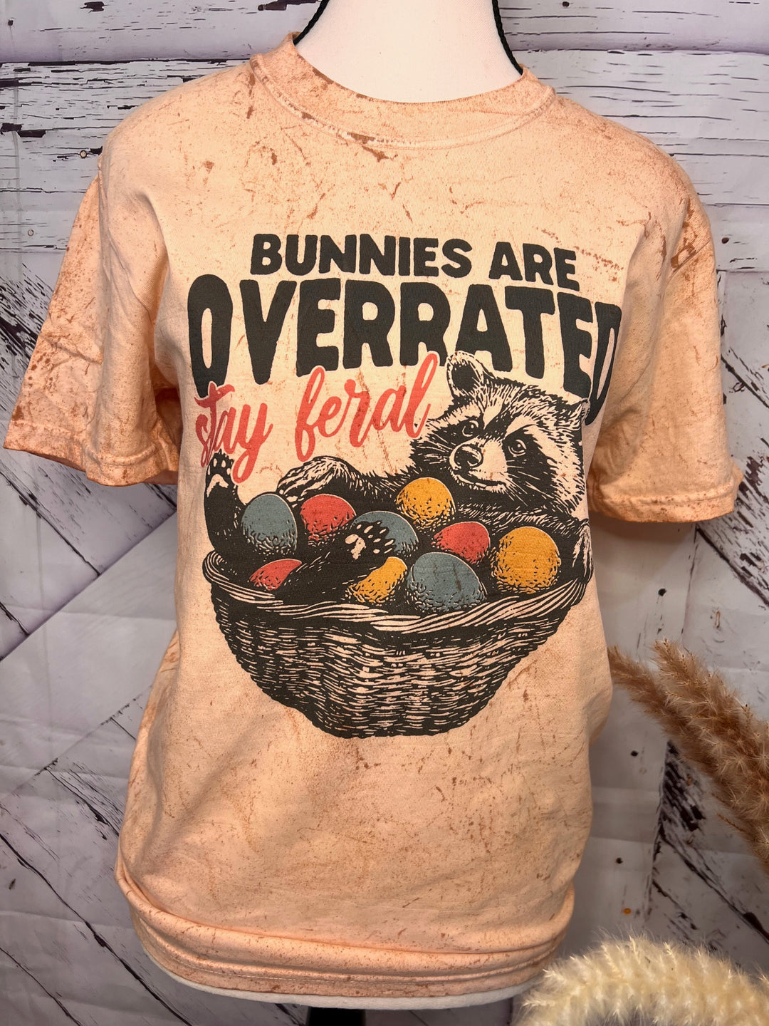 A peach-colored t-shirt with the text 'Bunnies are OVERRATED stay feral' and an image of a raccoon and a basket of colorful eggs.