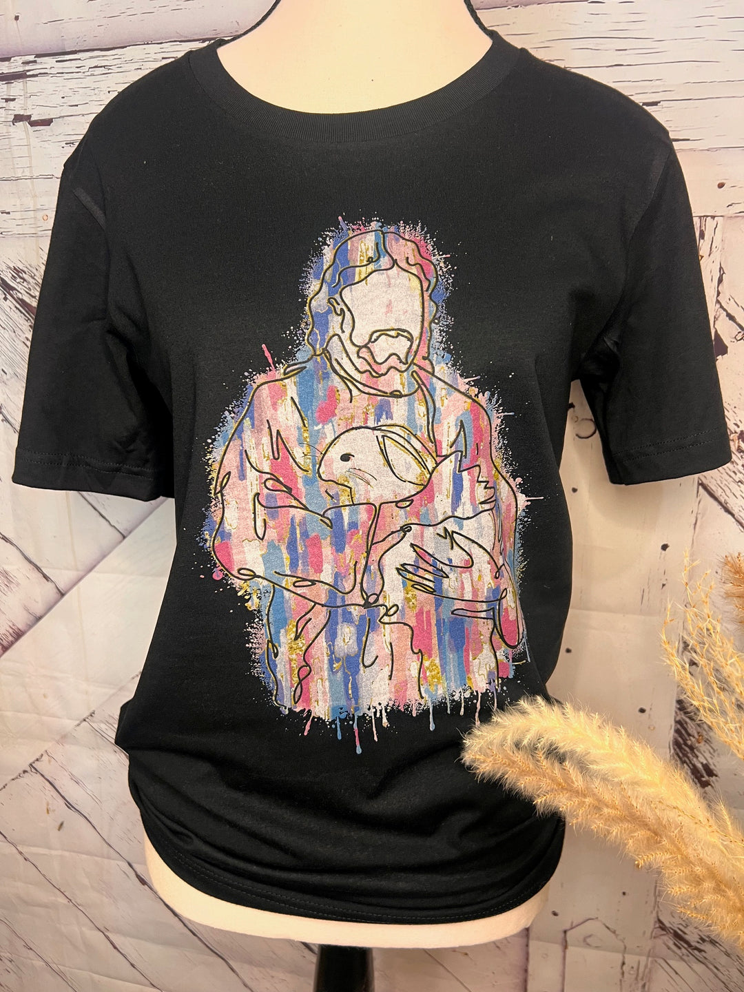 A black t-shirt featuring a graphic design of Jesus Christ holding a bunny with colorful, abstract patterns resembling paint splatters.