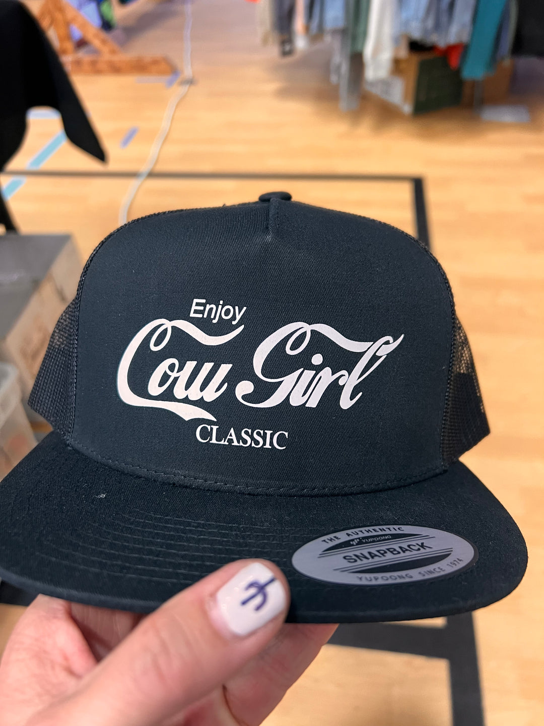 A black ballcap with 'Enjoy Cow Girl Classic' white text on it, resembling the Coca-Cola logo.