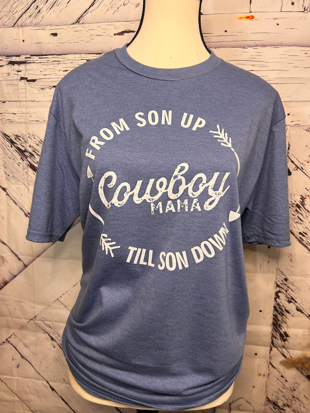 A blue Cowboy Mama graphic tee with a western-inspired design, featuring bold lettering and a circular design with arrows. The tee is displayed on a mannequin wearing a denim jacket and a beaded necklace.