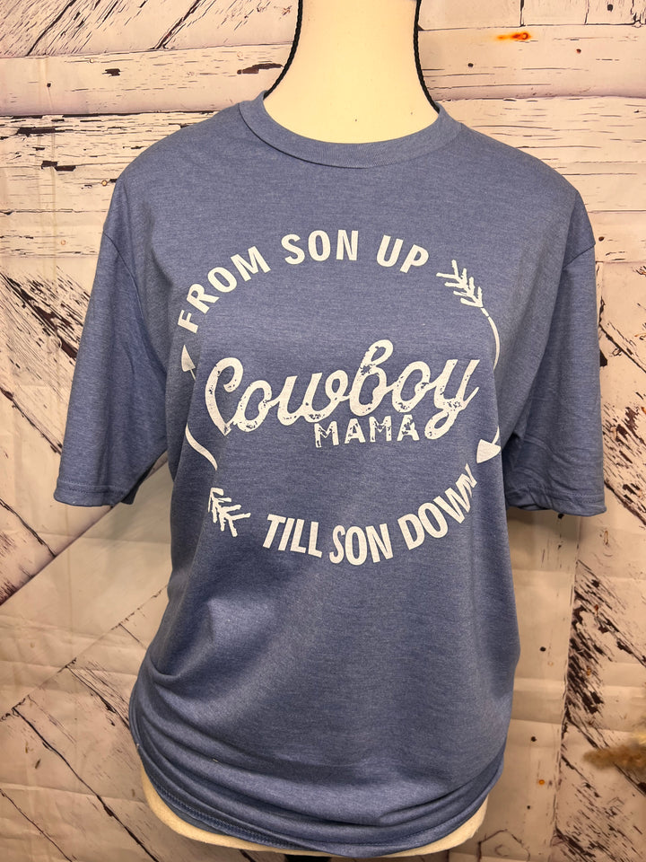 A blue Cowboy Mama graphic tee with a western-inspired design, featuring bold lettering and a circular design with arrows. The tee is displayed on a mannequin wearing a denim jacket and a beaded necklace.