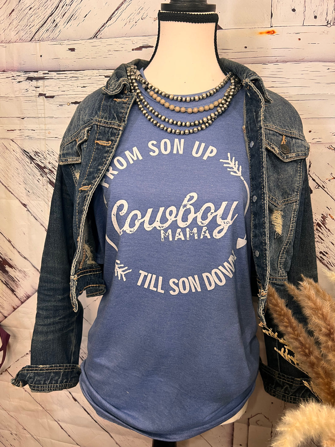 A blue Cowboy Mama graphic tee with a western-inspired design, featuring bold lettering and a circular design with arrows. The tee is displayed on a mannequin wearing a denim jacket and a beaded necklace.