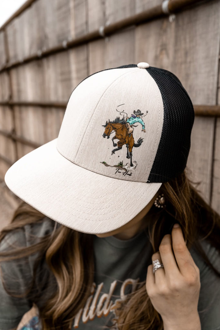 A beige and black trucker hat with a hand-drawn design of a cowgirl riding a bronc horse, held by a person against a wooden backdrop.