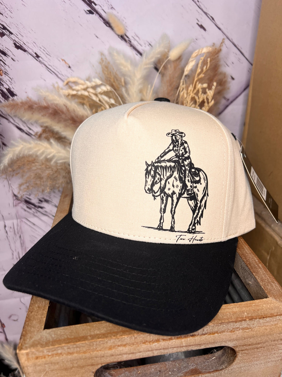 A beige and black ballcap with a hand-drawn outline of a cowgirl riding a horse on the front.
