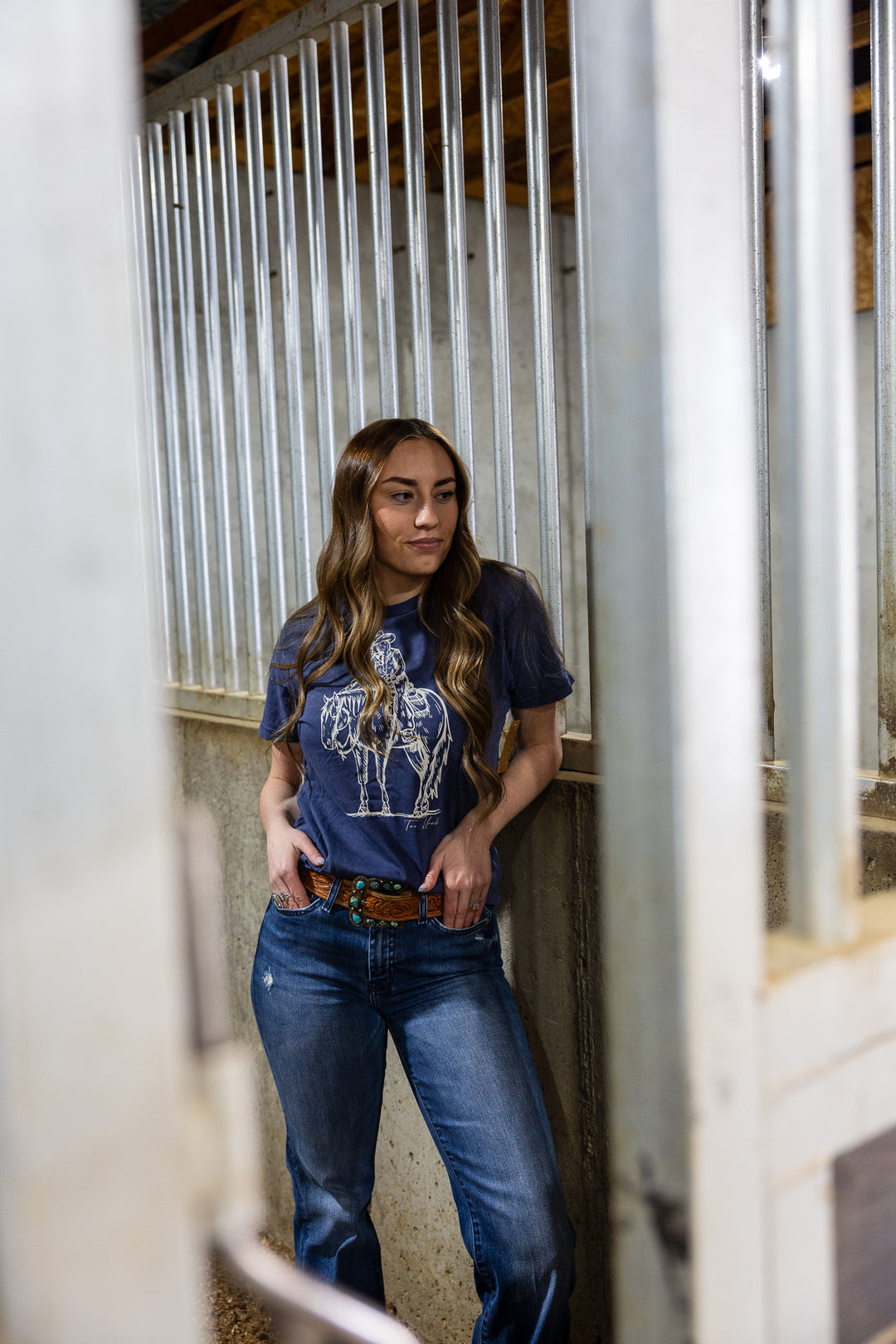 A woman standing in a stable wearing a navy t-shirt with a graphic of a cowgirl patting a horse's neck.