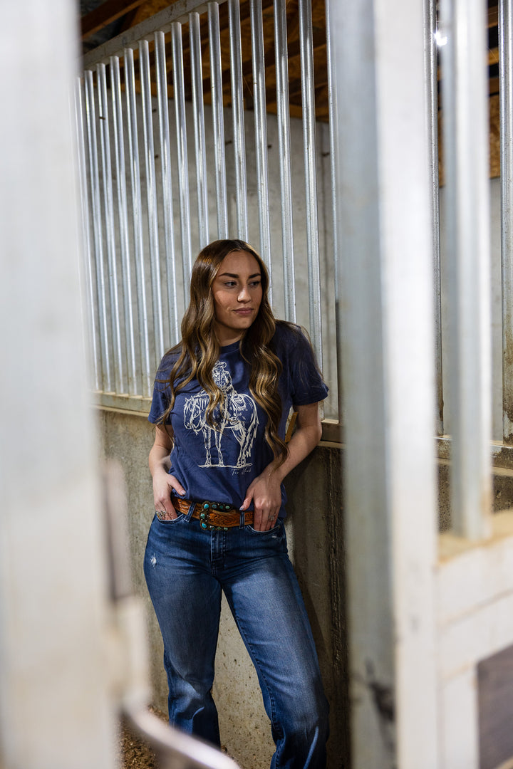 A woman standing in a stable wearing a navy t-shirt with a graphic of a cowgirl patting a horse&