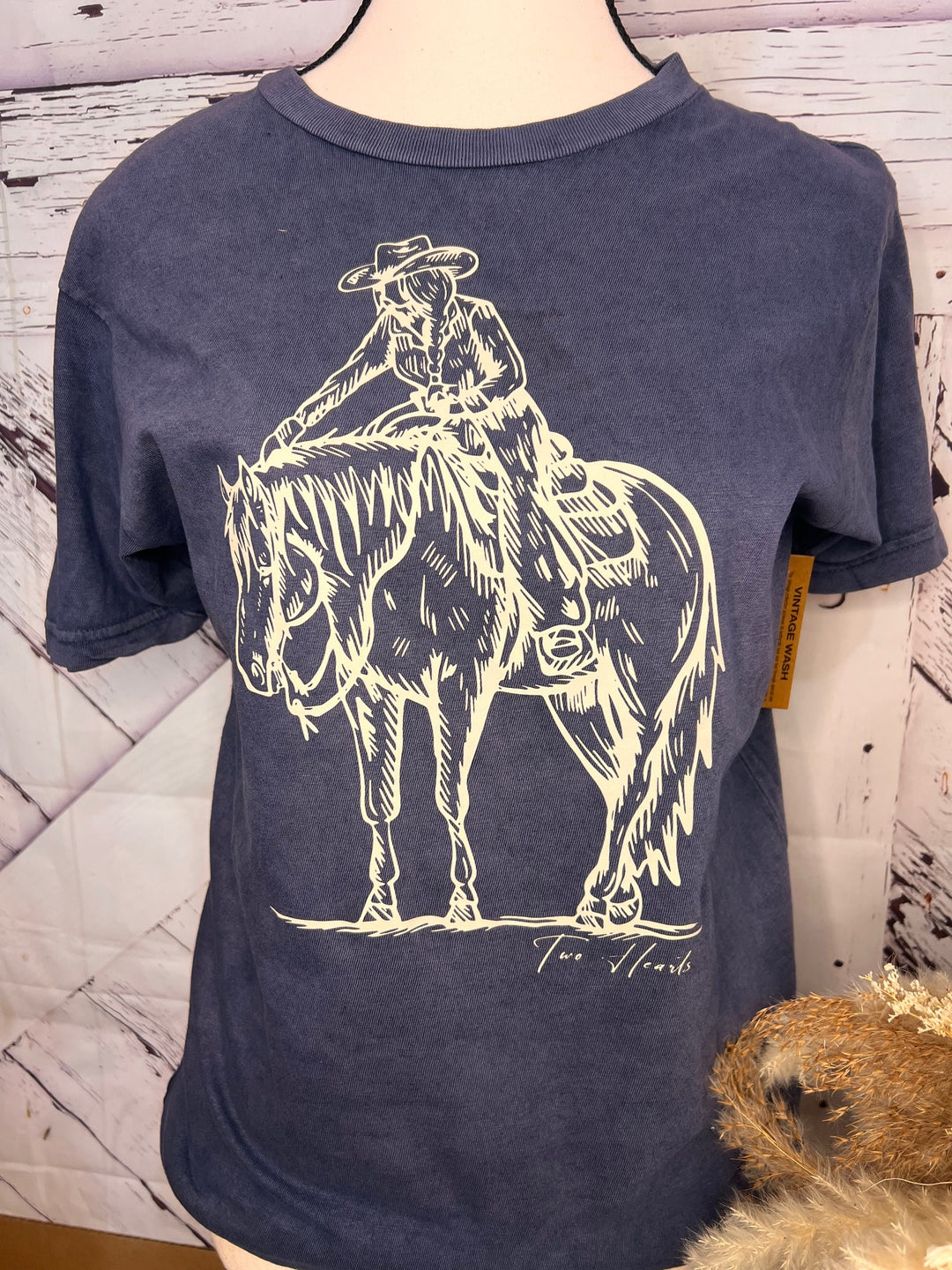 A woman standing in a stable wearing a navy t-shirt with a graphic of a cowgirl patting a horse&