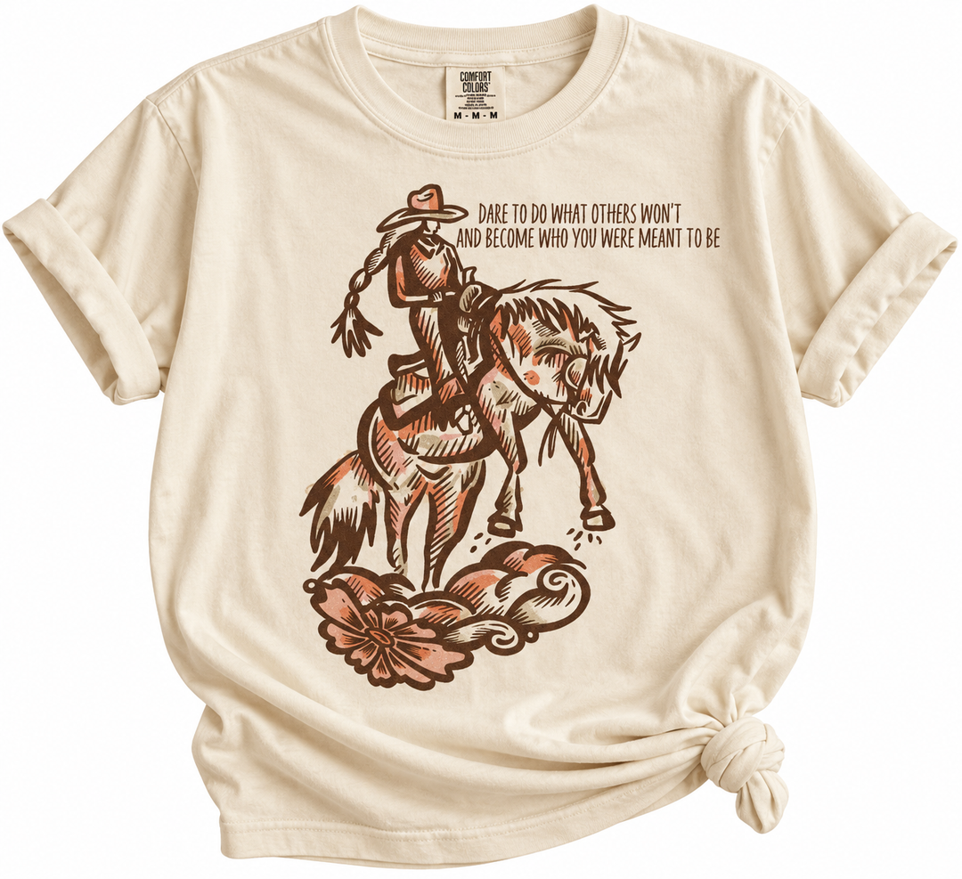 A cream-colored short-sleeve t-shirt with a rolled hem knot, featuring a brown graphic of a cowboy riding a bucking horse with the text 'DARE TO DO WHAT OTHERS WON'T AND BECOME WHO YOU WERE MEANT TO BE'.