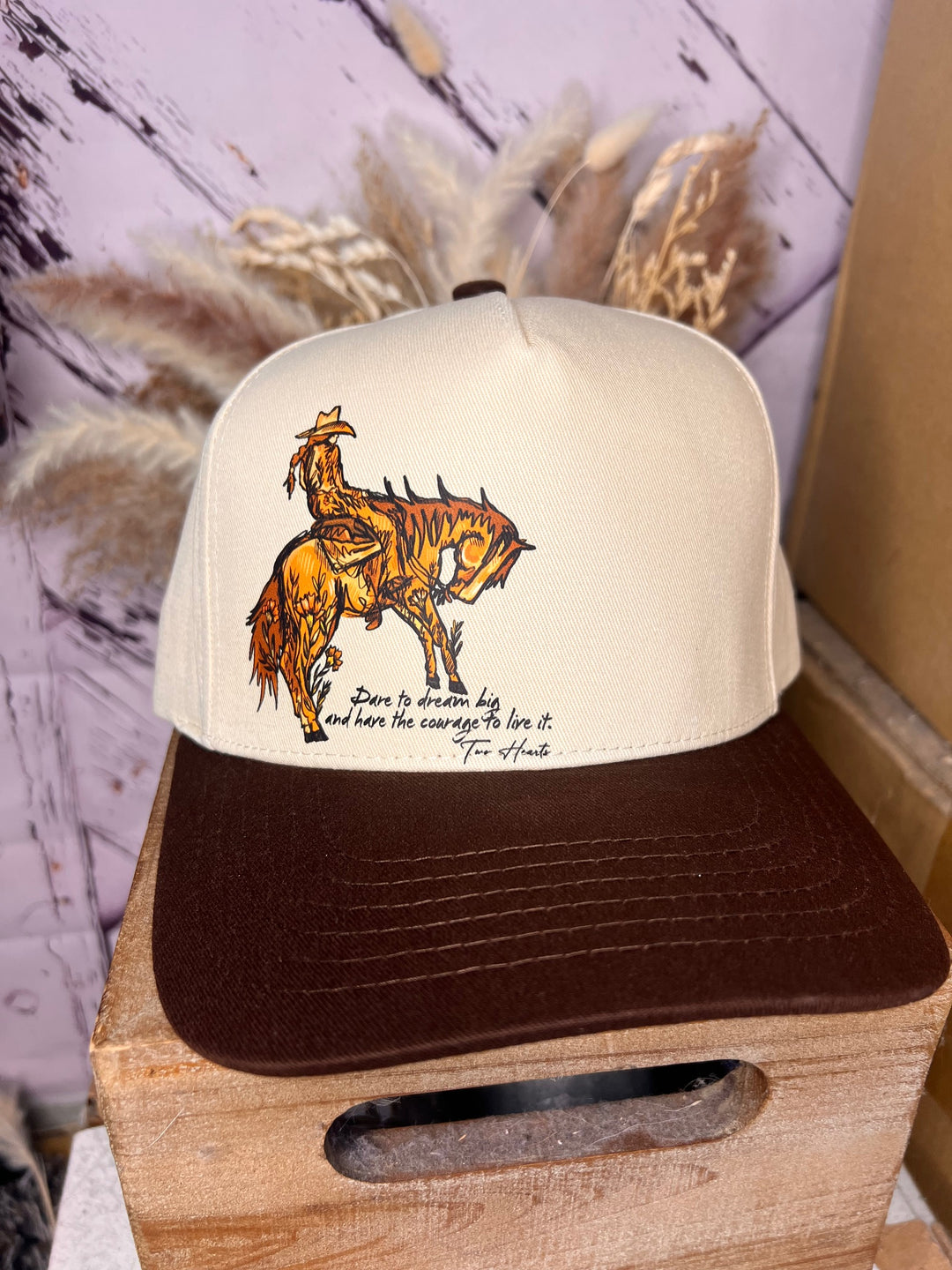 A beige and brown cowgirl ballcap with a graphic of a cowgirl riding a horse and the phrase 'Dare to dream big' printed on the front.