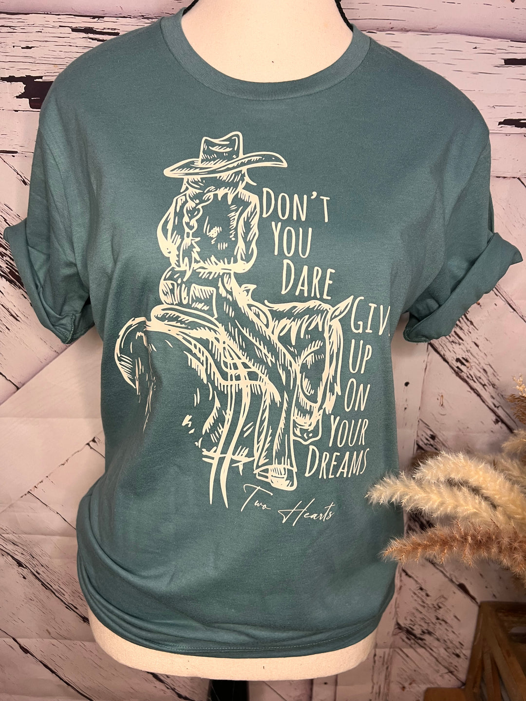 A green t-shirt featuring a white graphic of a cowgirl riding a horse, with the phrase 'Don't you dare give up on your dreams.'.