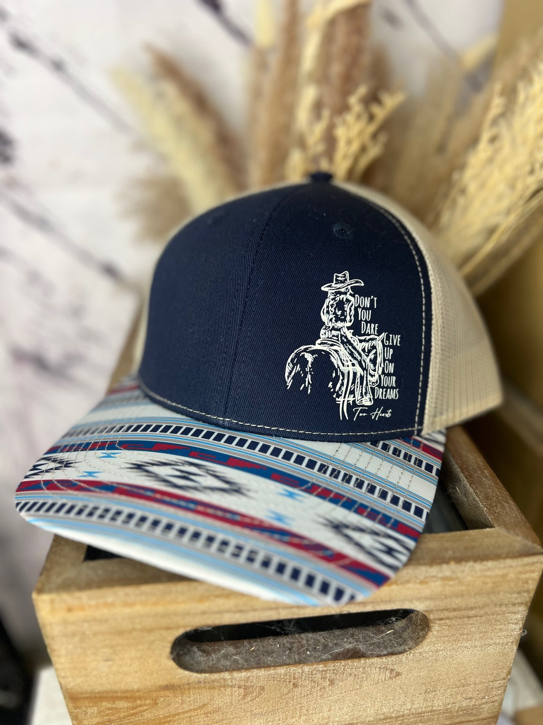 A navy and beige ballcap with a cowgirl riding a horse design, and a patterned brim, placed on a wooden surface.
