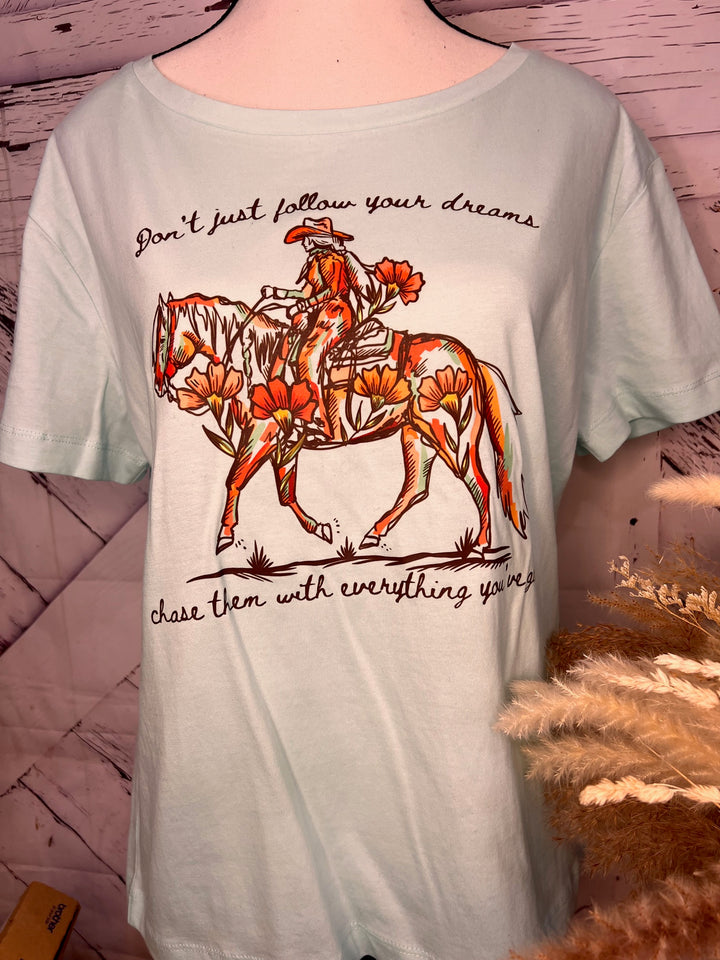 A woman standing outdoors wearing a pink graphic tee with a floral cowgirl design and the text &
