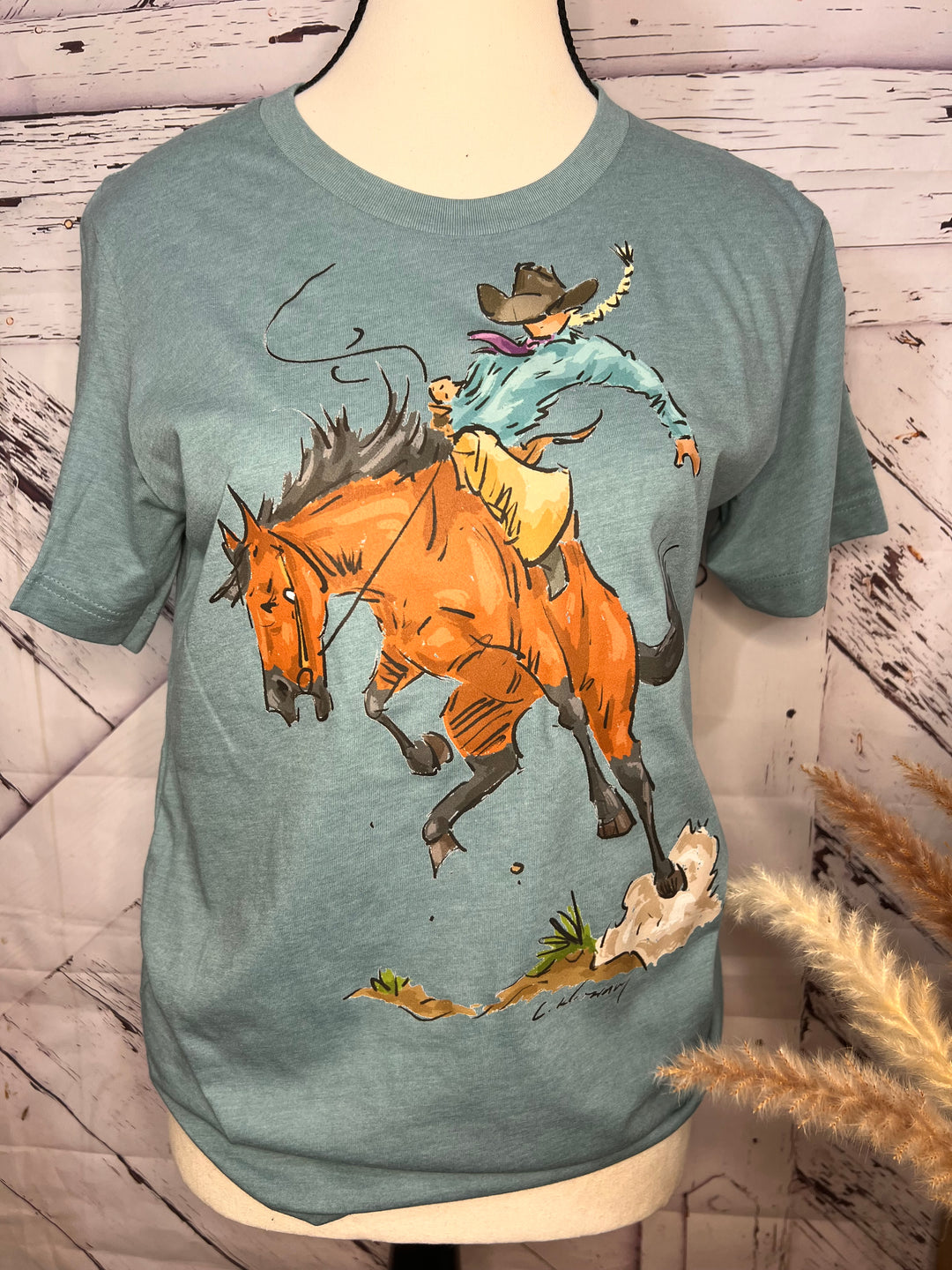 A green t-shirt with a hand-drawn graphic of a cowgirl riding a bucking bronc.