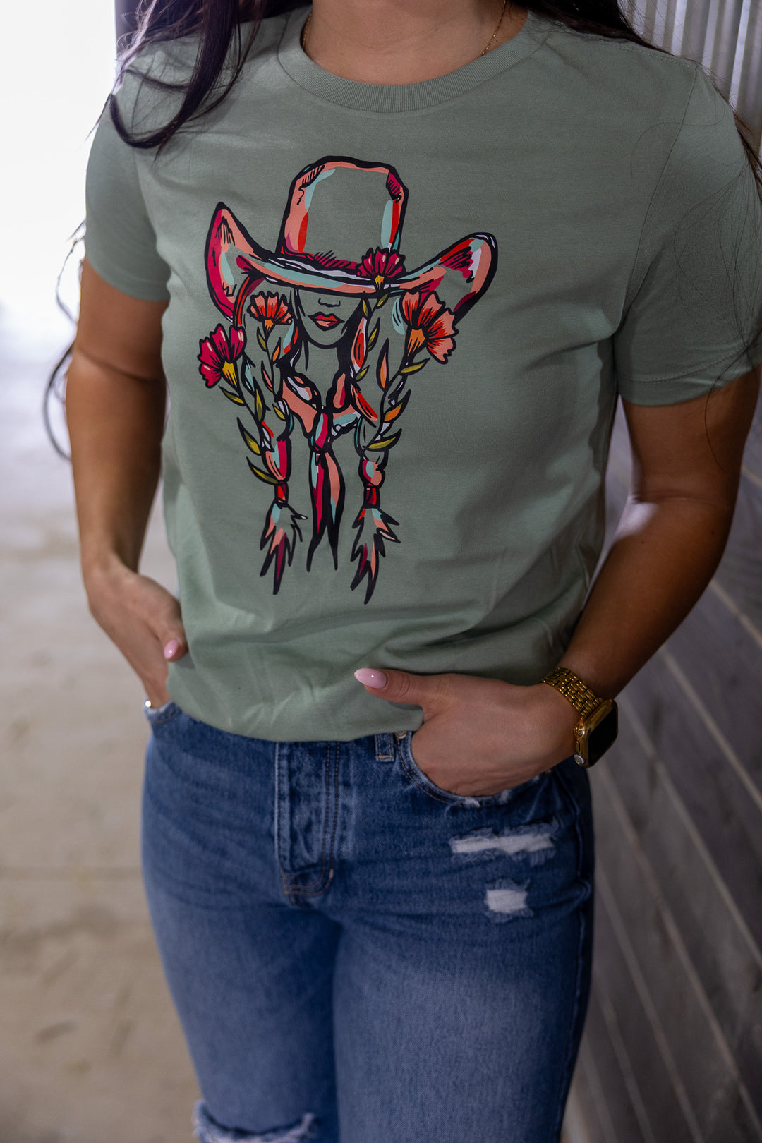 A person wearing a green graphic tee with a floral cowgirl design, featuring a cowgirl silhouette with a large cowboy hat and floral accents.