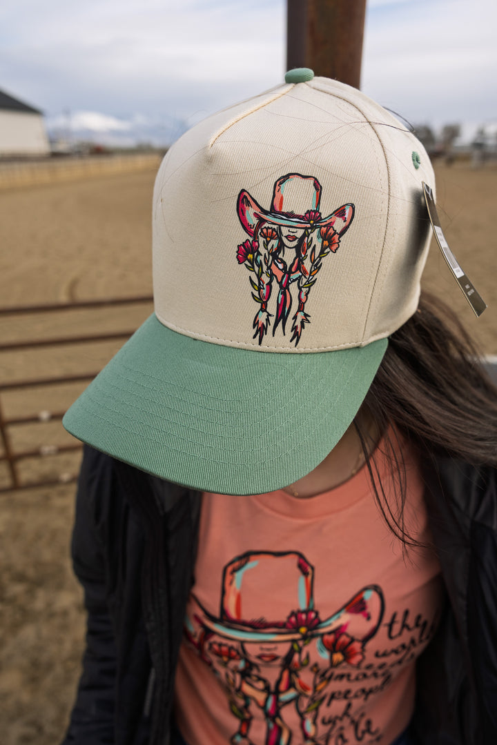 A floral cowgirl ballcap with a graphic of a cowgirl beneath a large cowboy hat on the front. The cap has a beige and green color scheme, and it is worn by a person with a pink shirt in the background.