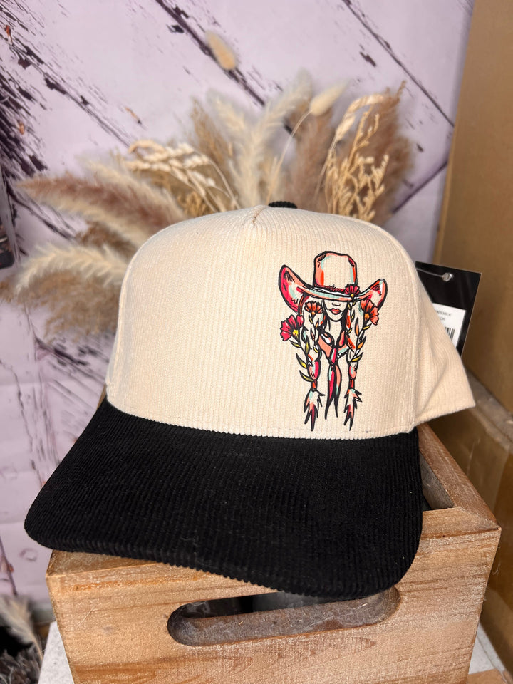 A floral cowgirl ballcap with a graphic of a cowgirl beneath a large cowboy hat on the front. The cap has a beige and green color scheme, and it is worn by a person with a pink shirt in the background.