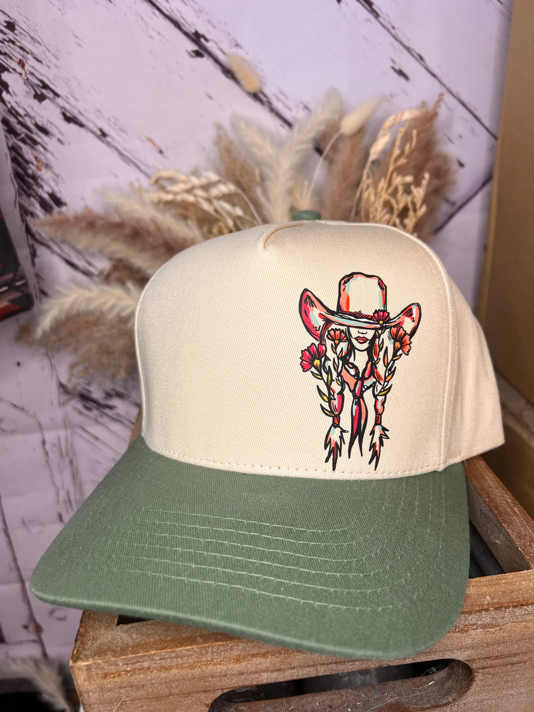 A floral cowgirl ballcap with a graphic of a cowgirl beneath a large cowboy hat on the front. The cap has a beige and green color scheme, and it is worn by a person with a pink shirt in the background.