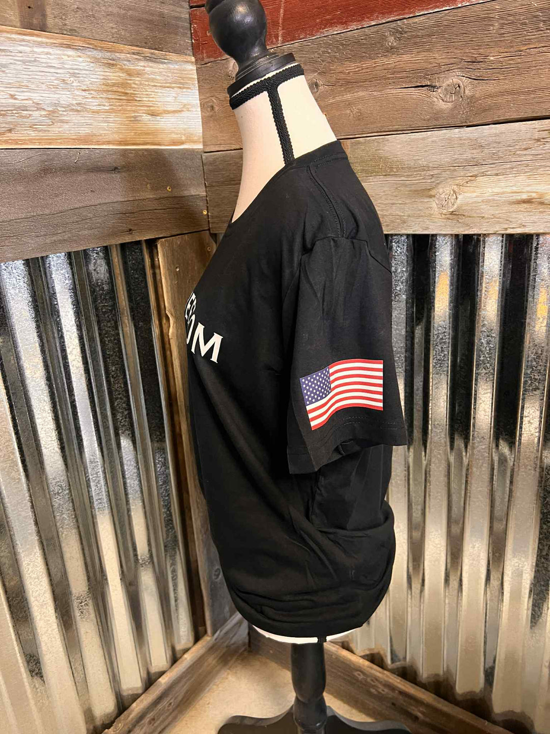 Black shirt with American flag patch on a mannequin against a rustic wooden wall.