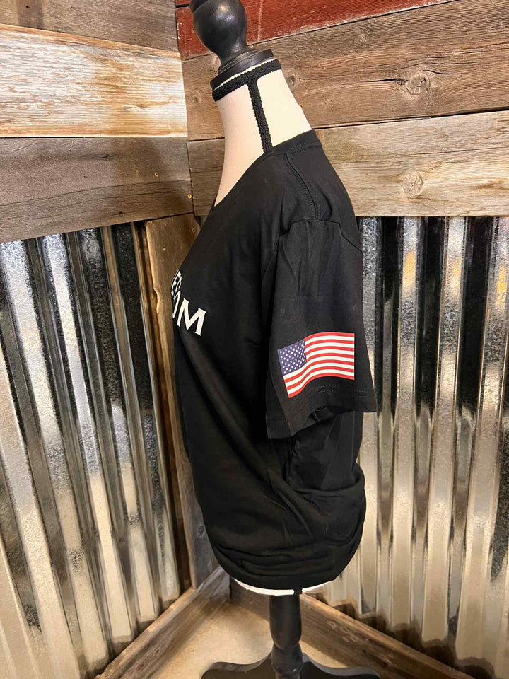 Black shirt with American flag patch on a mannequin against a rustic wooden wall.