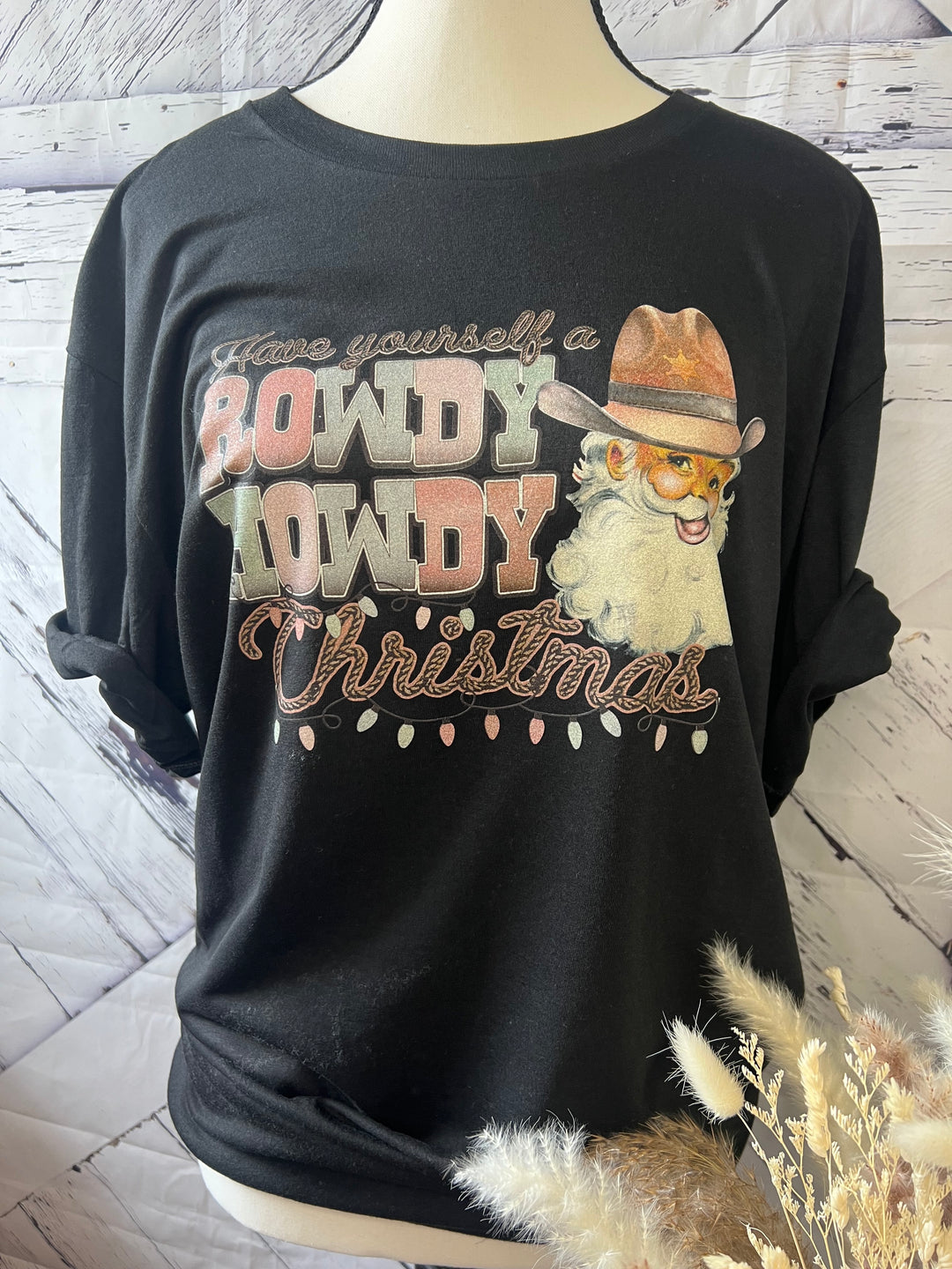 A black t-shirt with a Christmas themed graphic design featuring the text 'HOWDY' and an illustration of a cartoonish Santa Claus wearing a cowboy hat.