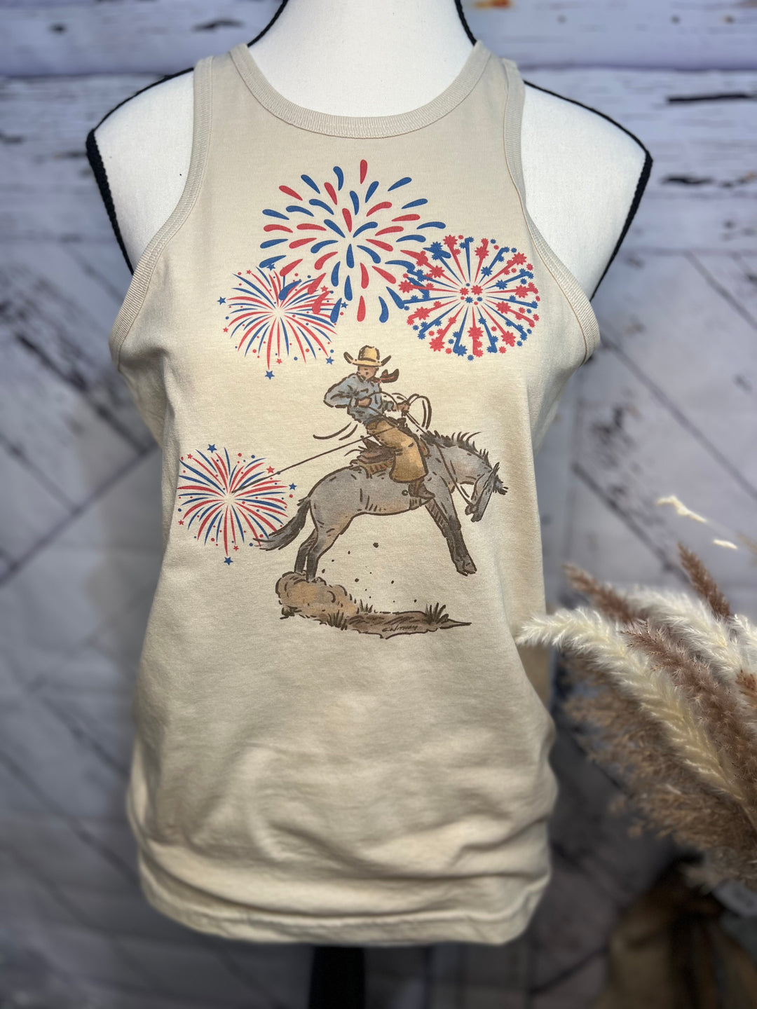 A beige tank top featuring a graphic print of a cowboy on a bucking horse with a background of fireworks.