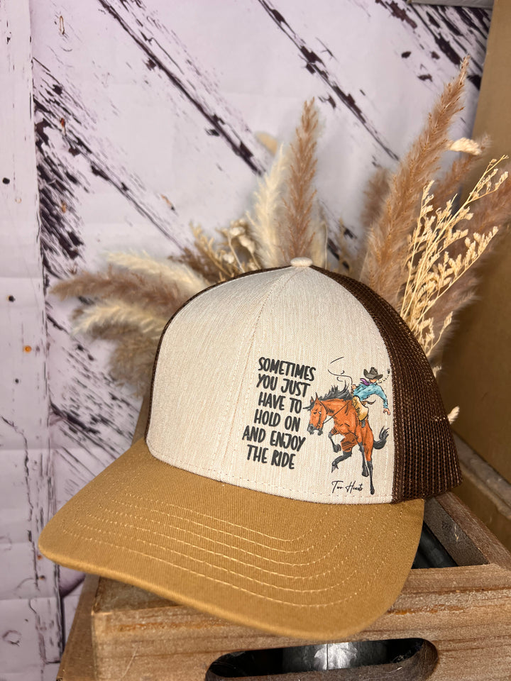 A person wearing a beige and green trucker hat with a cowgirl riding a bronc illustration on it.