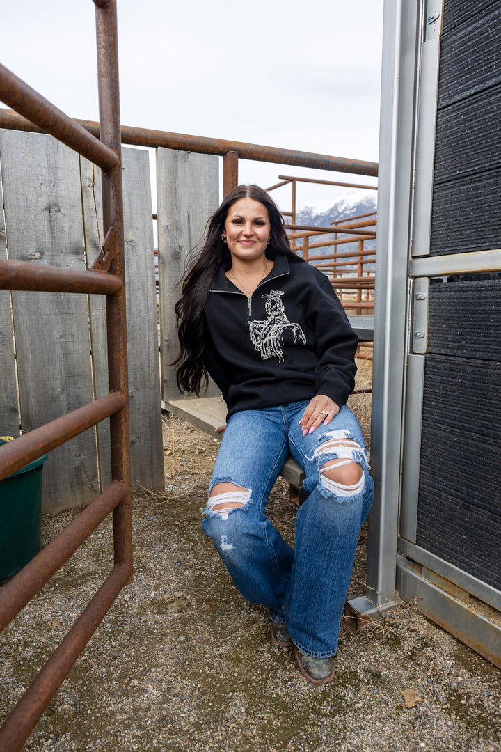 A woman sitting on a gate post wearing a black quarter zip sweatshirt with a white line-drawn cowgirl design and blue ripped jeans.