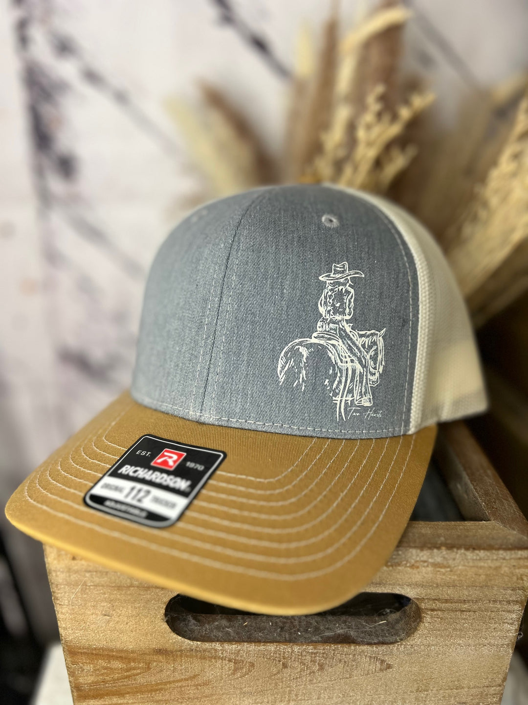 A ballcap with a minimalist white outline of a cowgirl riding a horse on a grey background with a yellow bill, displayed on a wooden surface.