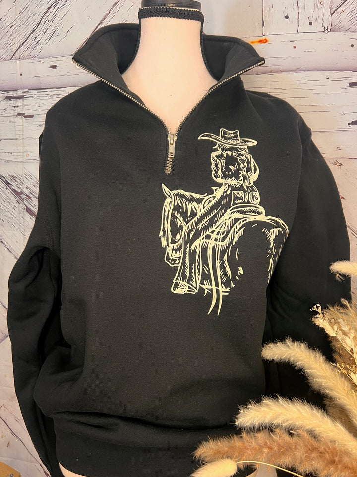 A woman sitting on a gate post wearing a black quarter zip sweatshirt with a white line-drawn cowgirl design and blue ripped jeans.
