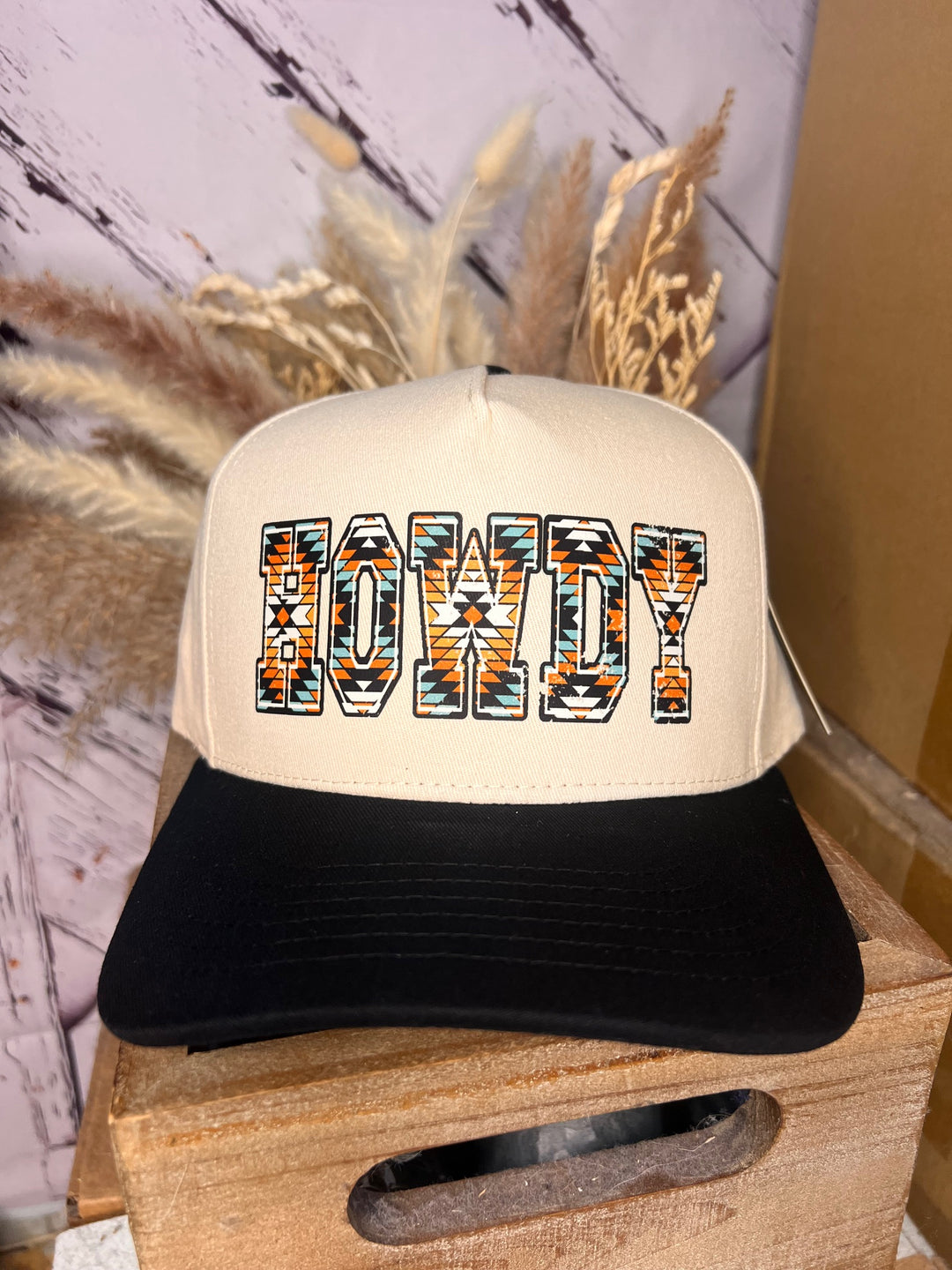 A ballcap with 'HOWDY' written in a Western Aztec font, featuring a beige crown and a black visor.