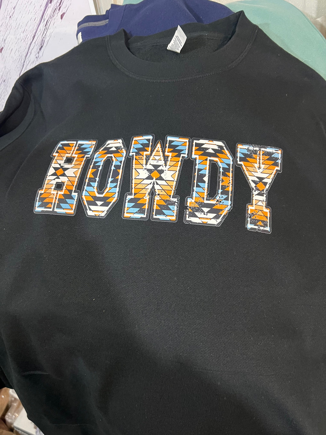 A black crewneck sweatshirt with the word 'HOWDY' written in a vibrant, Aztec-inspired geometric pattern.