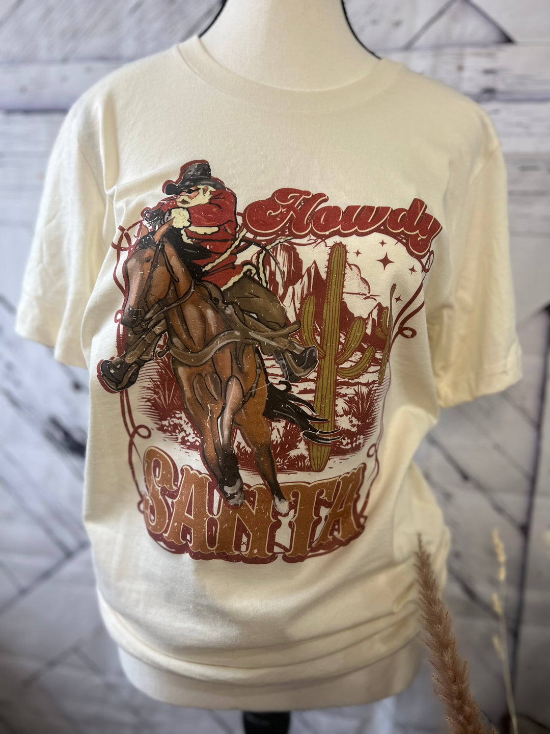 A white crew neck t-shirt with a graphic print of Santa Claus riding a horse, with the text 'HOWDY SANTA' and a Christmas tree in the background.