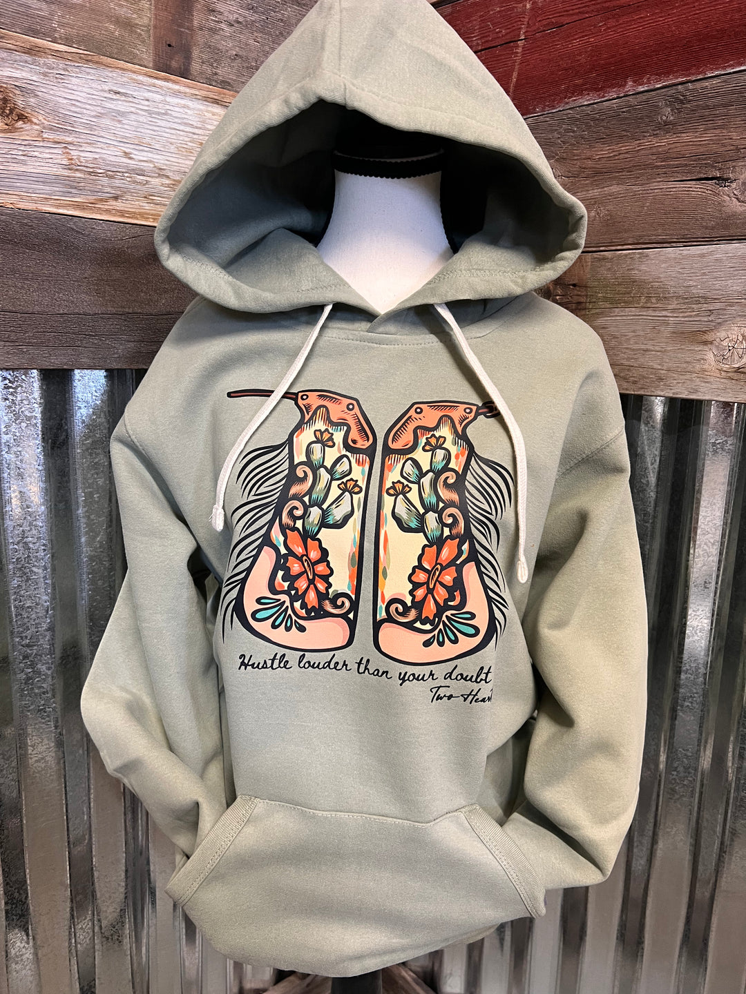 A sage green pullover hoodie displayed on a mannequin torso, featuring a large graphic of two cowboy boots with floral designs and the text &