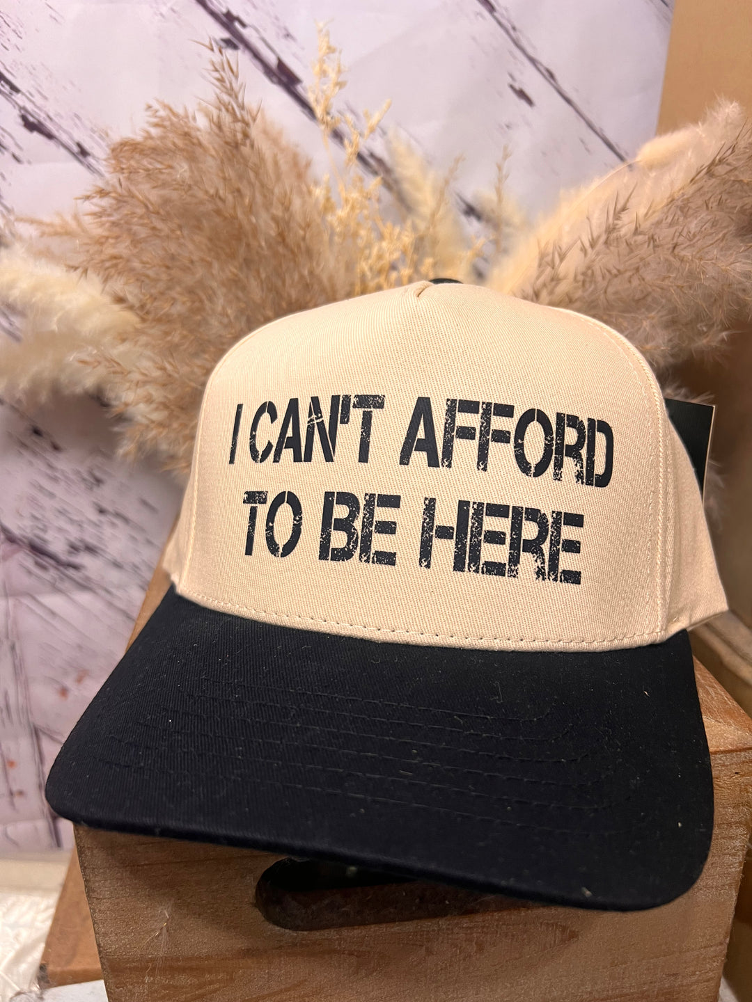 A khaki and black ballcap with the text 'I can't afford to be here' printed in the front.