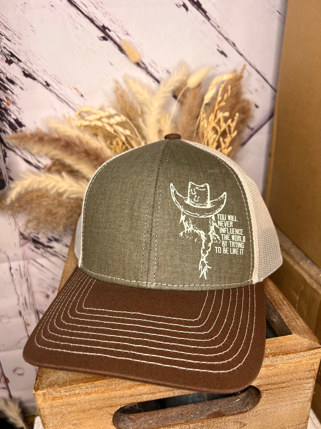 A beige, brown, and blue trucker ballcap with a cowgirl design and text on the front panel, displayed on a wooden surface with decorative elements in the background.