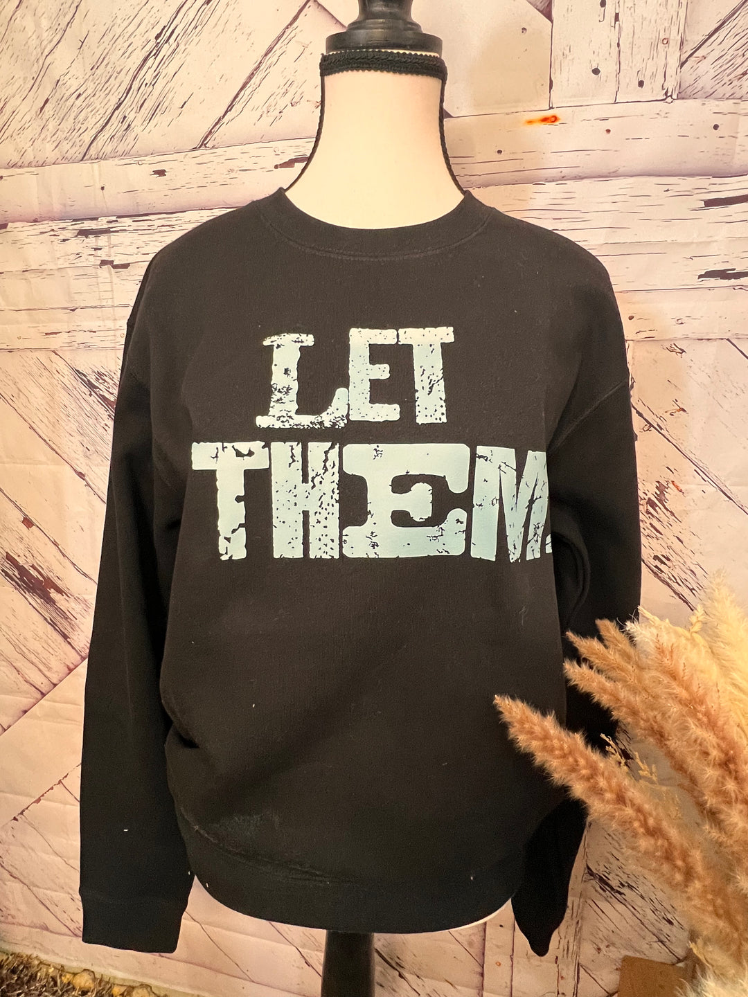 A black crewneck sweatshirt with the phrase 'LET THEM' written in distressed turquoise lettering on the front.