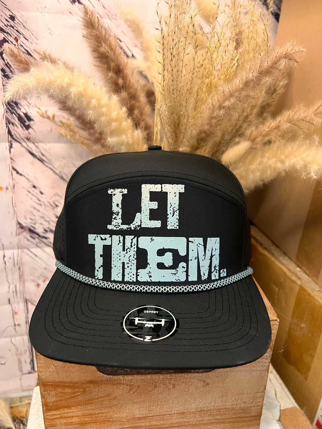 A black ballcap with distressed turquoise 'LET THEM' lettering on the front and a matching turquoise rope detail on the brim, displayed on a wooden surface with decorative dry plants in the background.