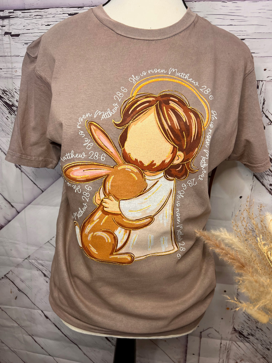 A graphic t-shirt with a design of a faceless Jesus Christ embracing an Easter bunny, with the text 'He is Risen' from Matthew 28:6.