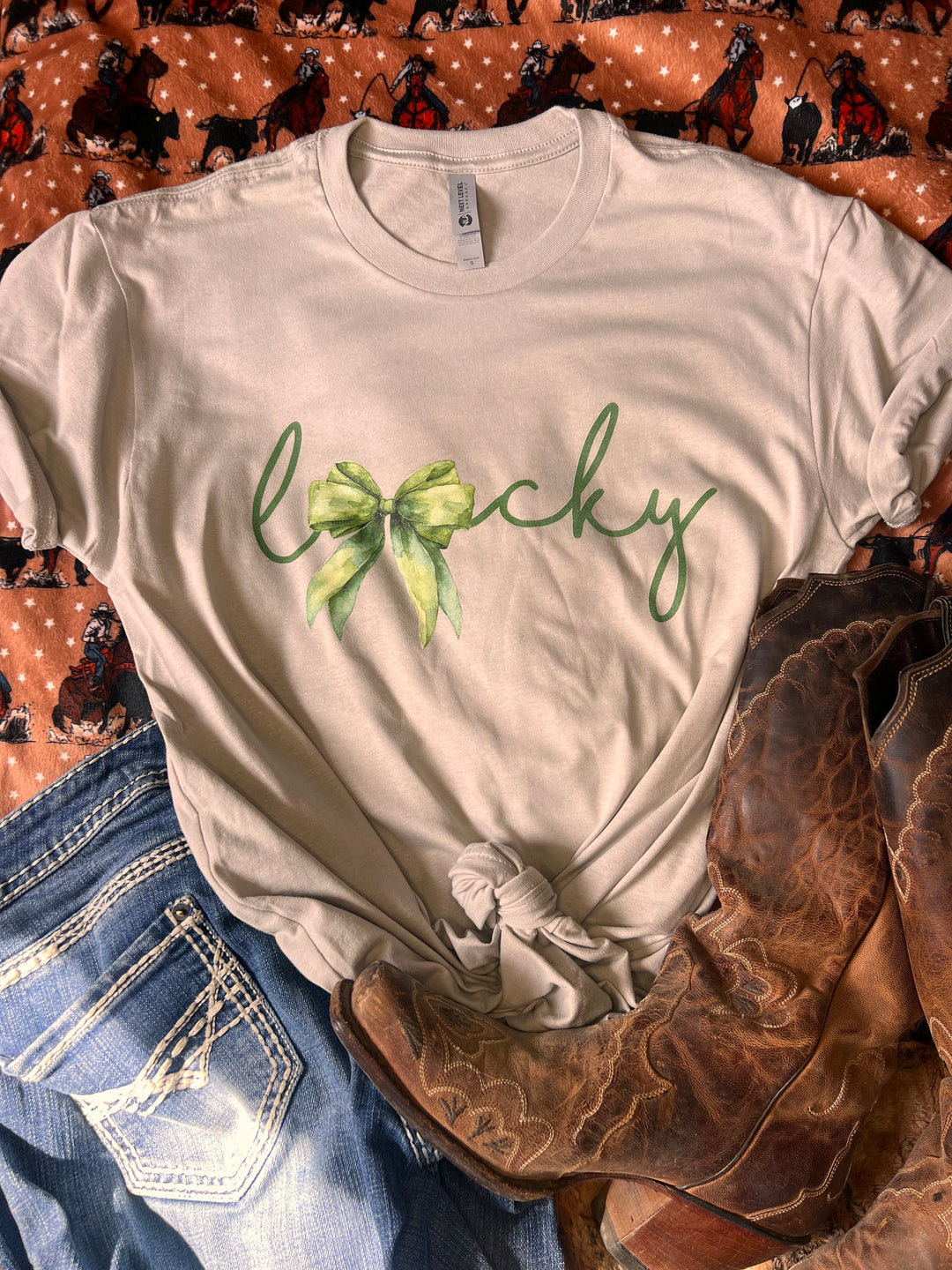 A t-shirt with the word 'Lucky' printed in green, where the 'u' is replaced with a western-style bow, laid out flat with a patterned background.