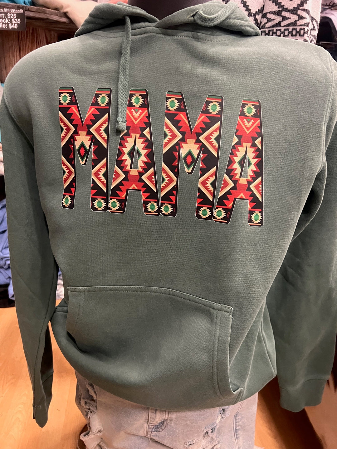 A forest green hoodie with the word 'MAMA' written in stylized Aztec-patterned letters on the back.